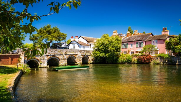 A river with a stone bridge and a green boat. There are buildings with red and white facades along the riverbank.