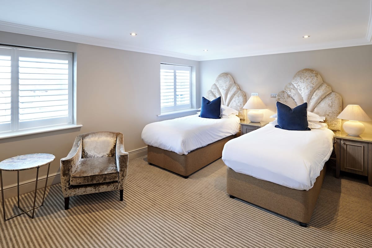 A hotel room with two beds, a small round table, a vintage armchair, and two bedside lamps.