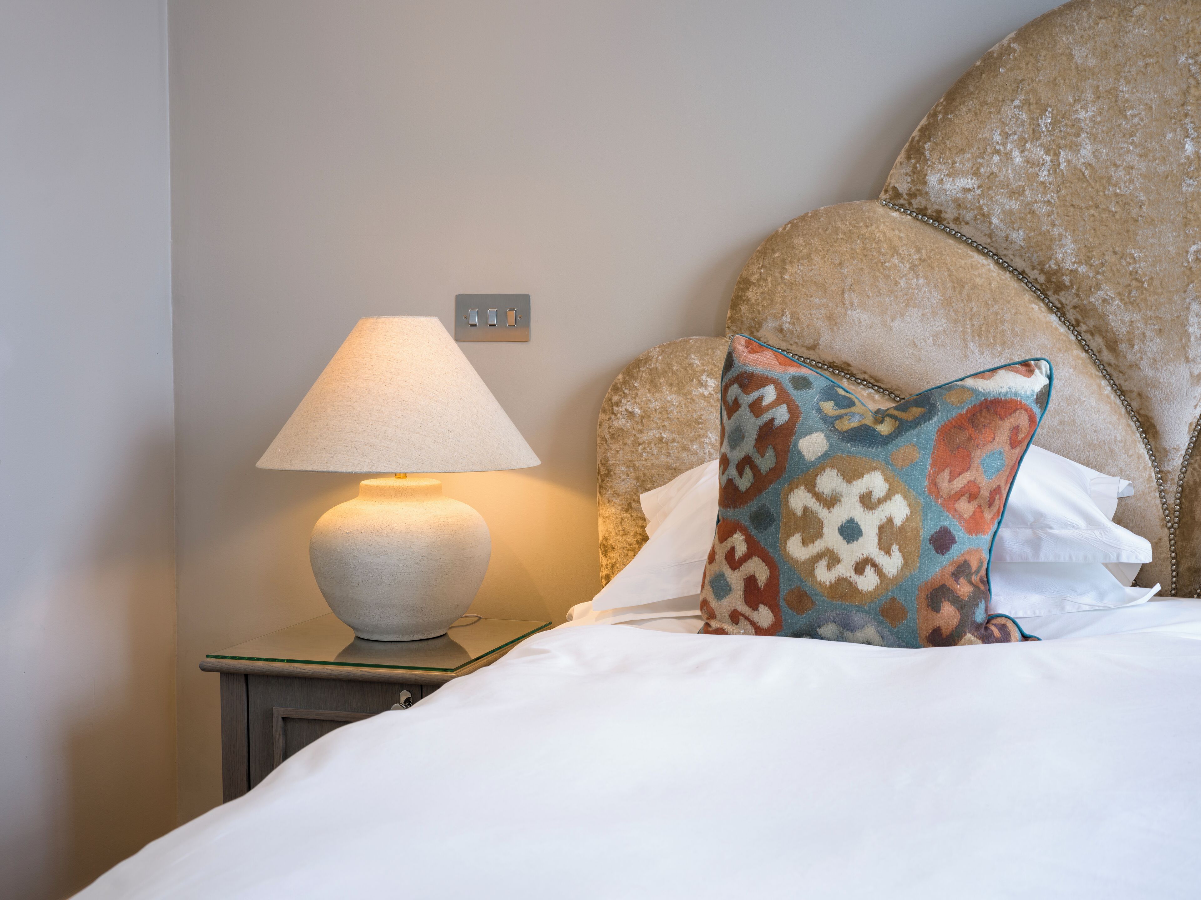 A neatly made bed with white linens, a patterned pillow, a table lamp, and a headboard.