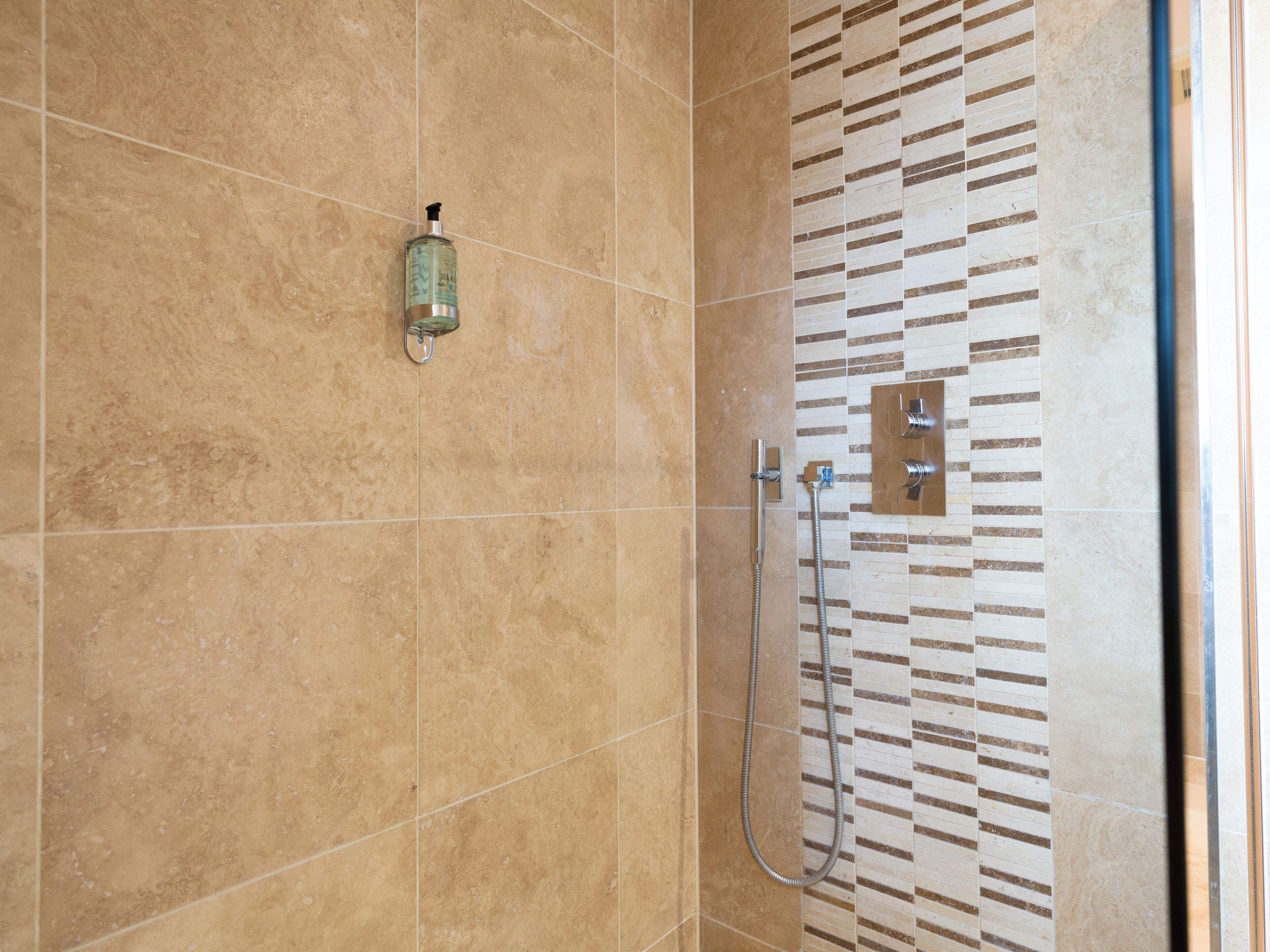 Une salle de bain avec des carreaux de couleur beige, une douche équipée d’un pommeau de douche amovible et un distributeur de savon fixé au mur.