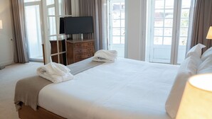 A neatly made bed with white linens and towels, a wooden dresser with a television, and a view through a glass door. - Hotel Toural (Guimarães)