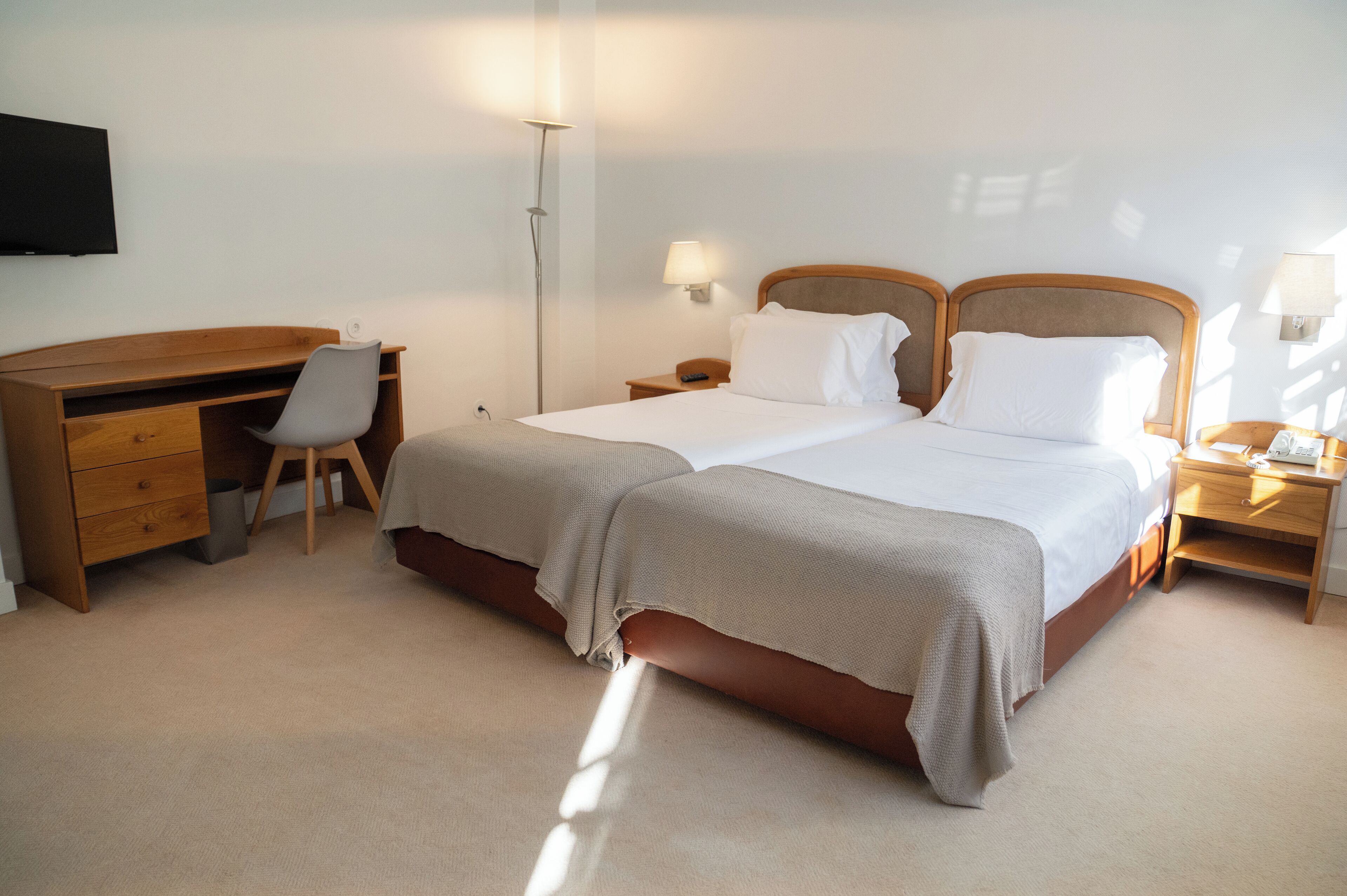 A hotel room with a double bed, a desk with a chair, a TV mounted on the wall, and a bedside table with a lamp.