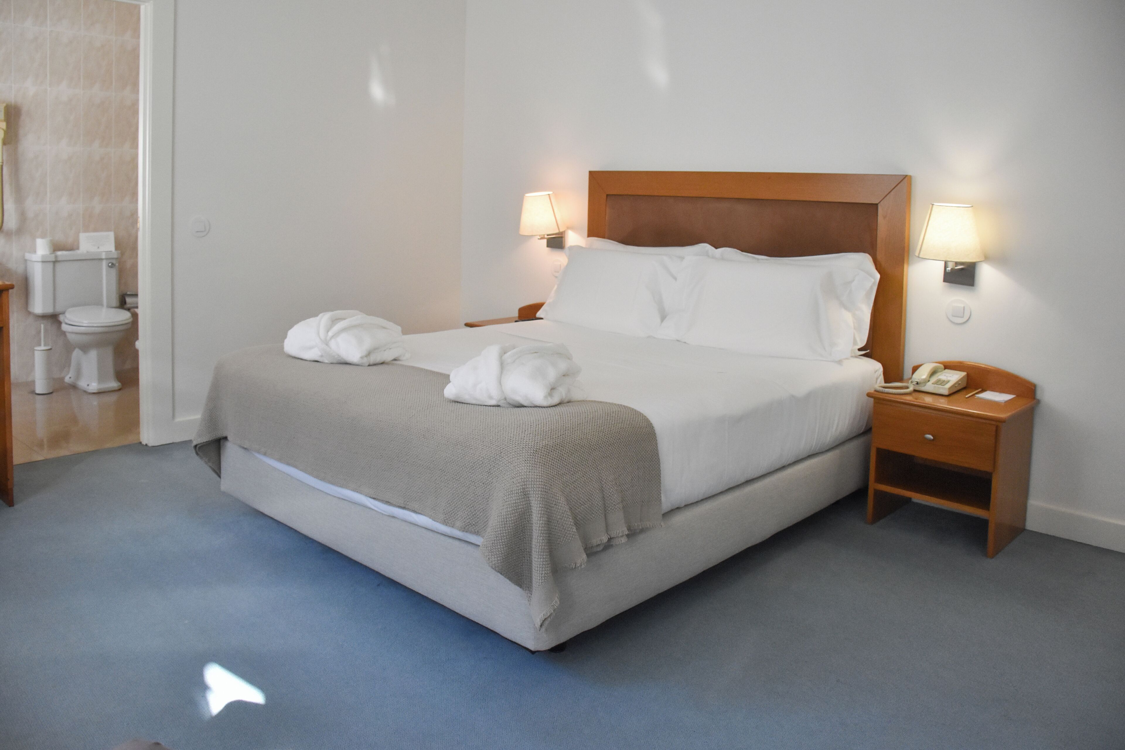 Una cama bien hecha con sábanas blancas y un cabecero de madera, dos lámparas de noche, un teléfono junto a la cama y una mesita de noche pequeña.