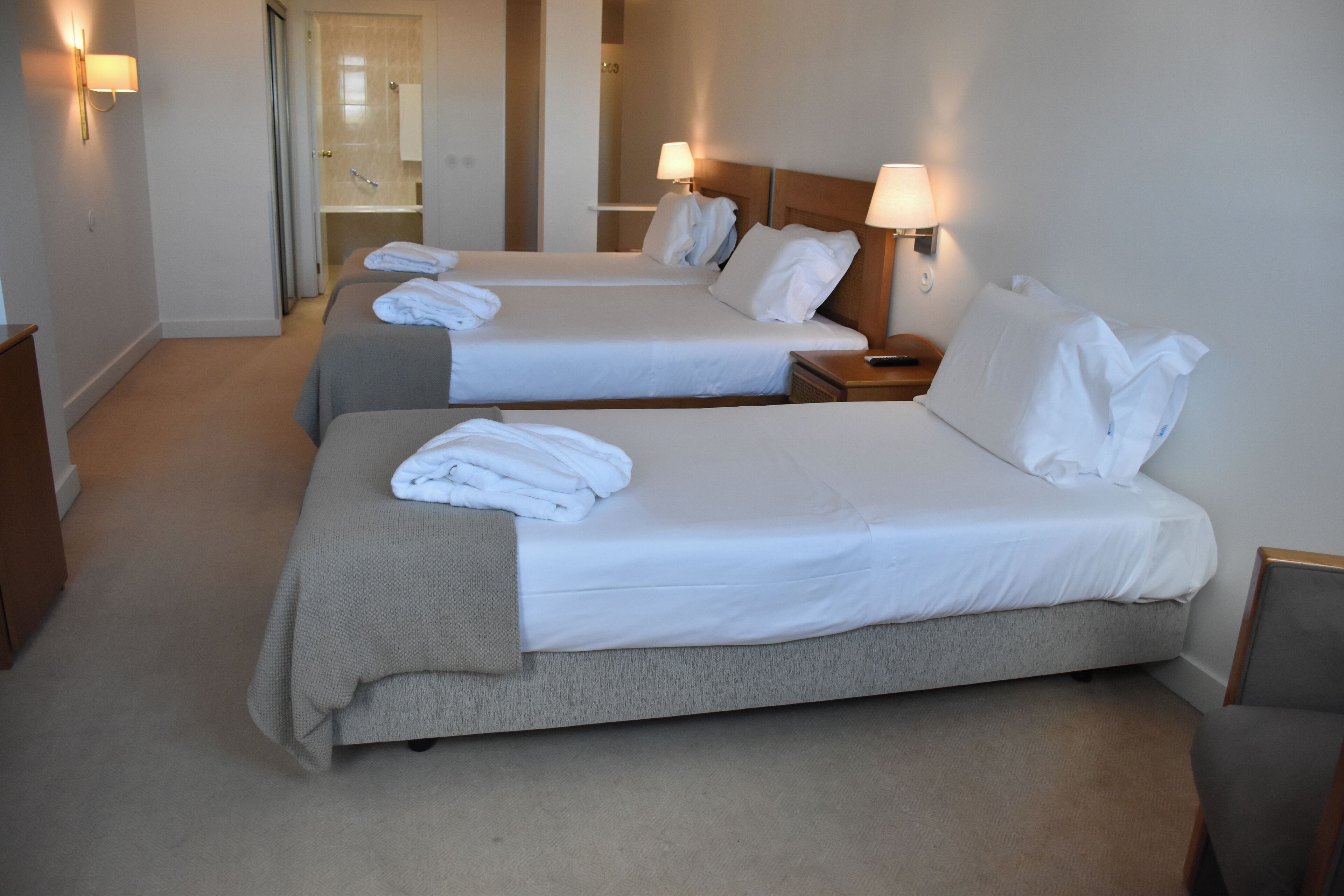 A hotel room with two beds, each with a pillow and a folded towel. There is a nightstand with a lamp and a wooden chair.