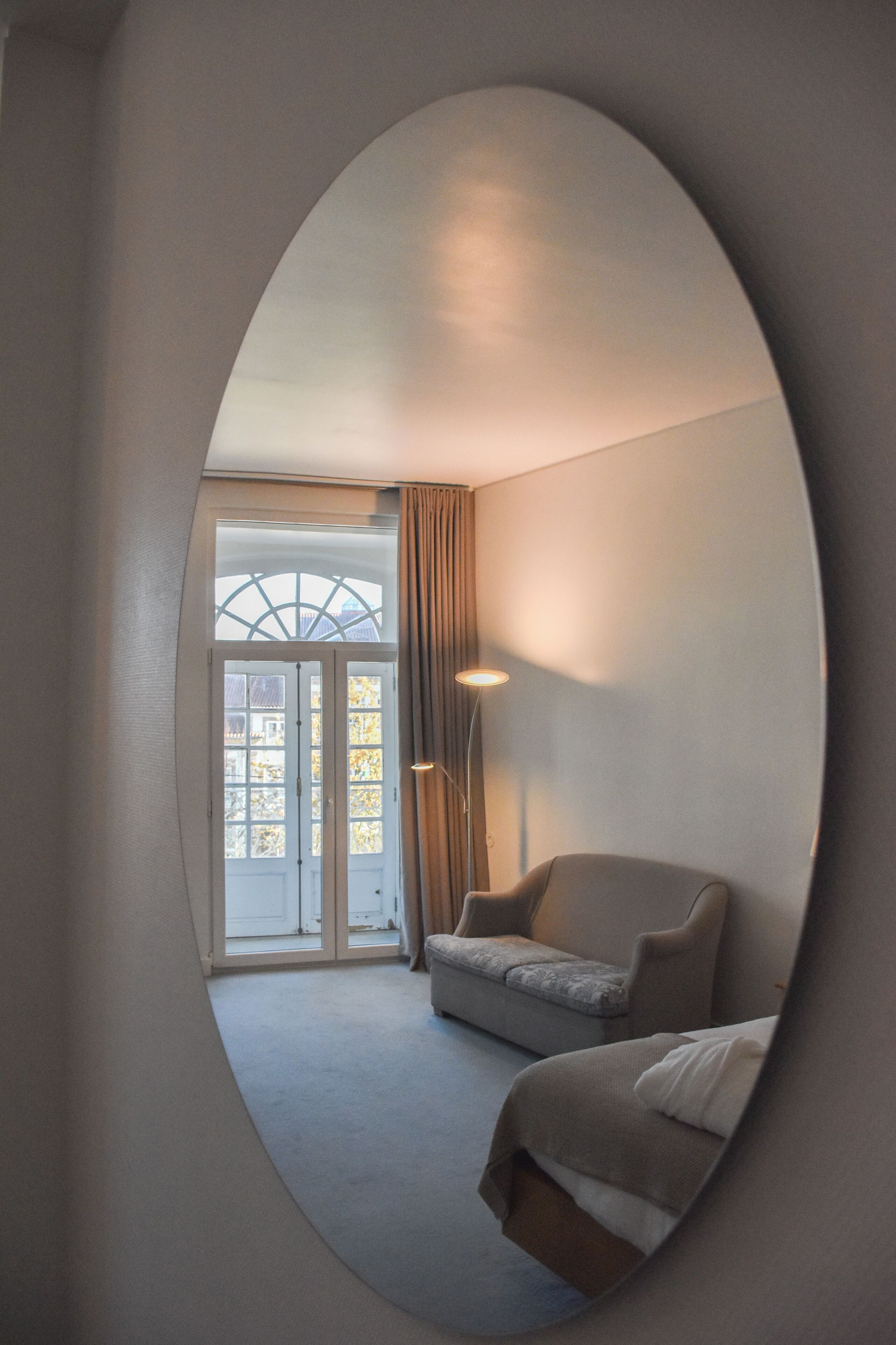 Habitación de hotel con un espejo ovalado grande, un sofá, una cama, una lámpara y vistas al exterior a través de un ventanal.