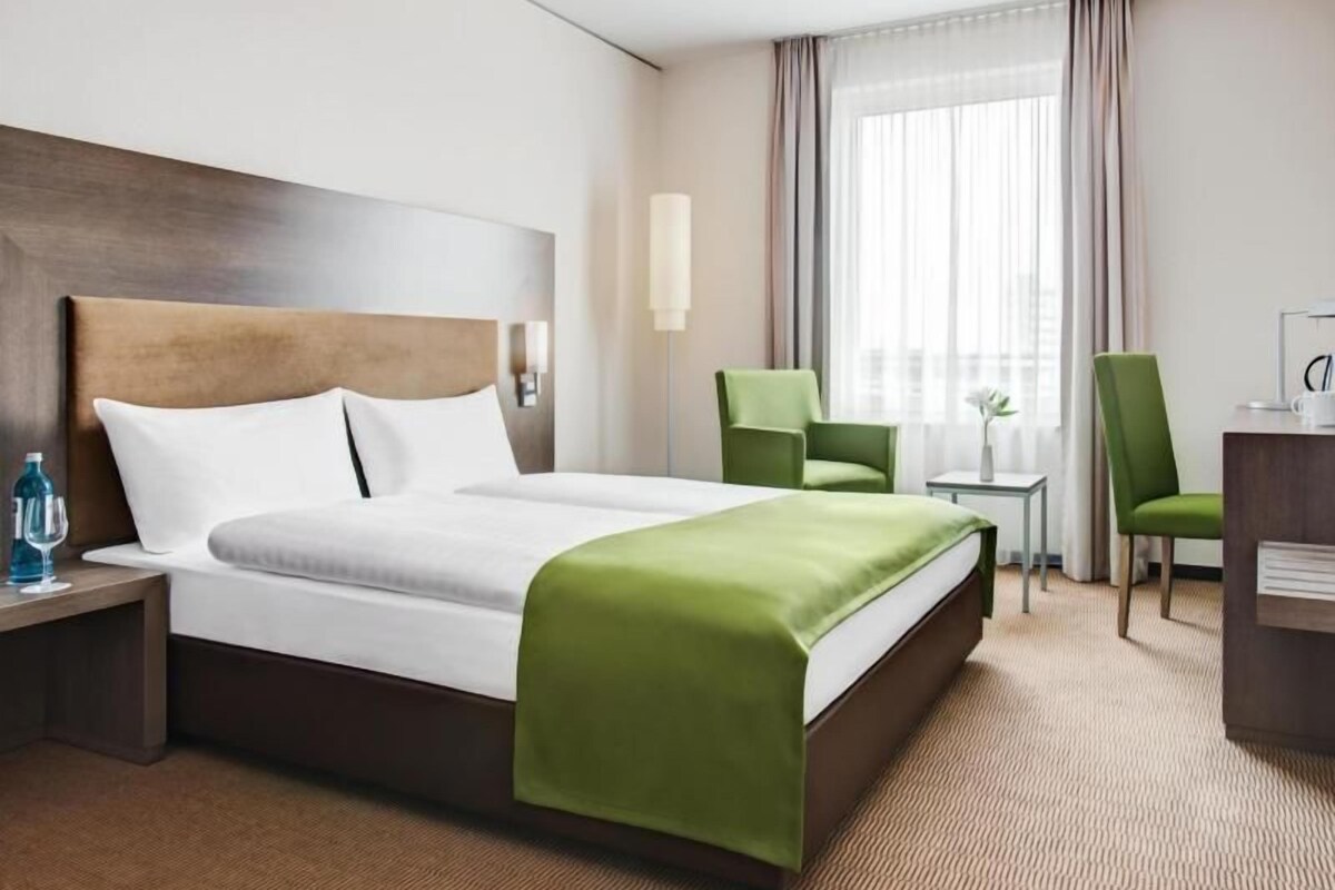 Una habitación de hotel con una cama grande, un escritorio, dos sillas, una mesita de noche y una ventana con cortinas.
