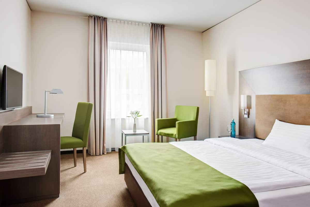 A hotel room with a bed, a desk, a chair, a TV, a lamp, and a window with curtains.