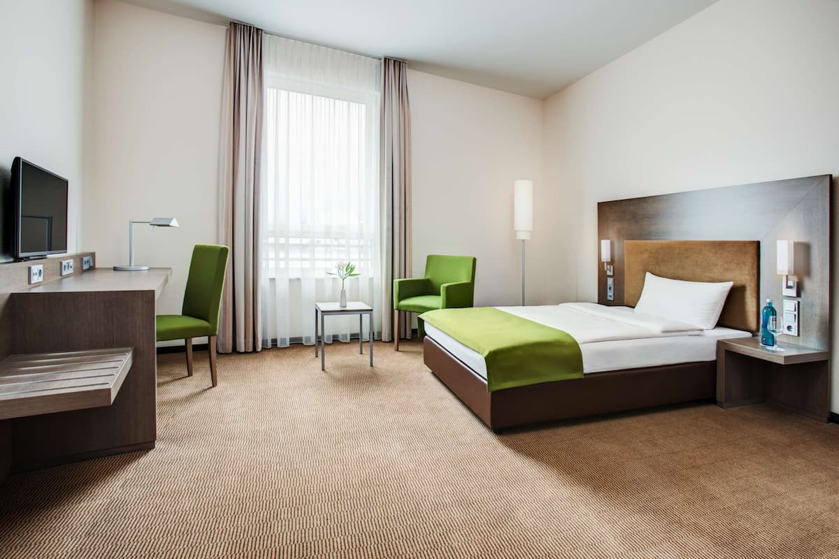 A hotel room with a bed, two chairs, a desk with a TV, a lamp, and a window with curtains.