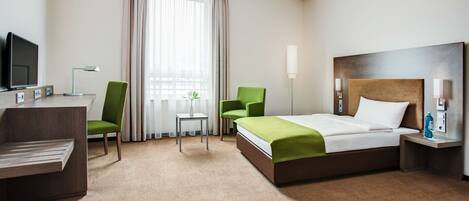 Quarto de hotel com cama, duas cadeiras, escrivaninha com televisão, abajur e janela com cortinas.