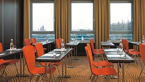 Un salón de conferencias con mesas y sillas, ventanas amplias con cortinas y vista a un paisaje urbano.
