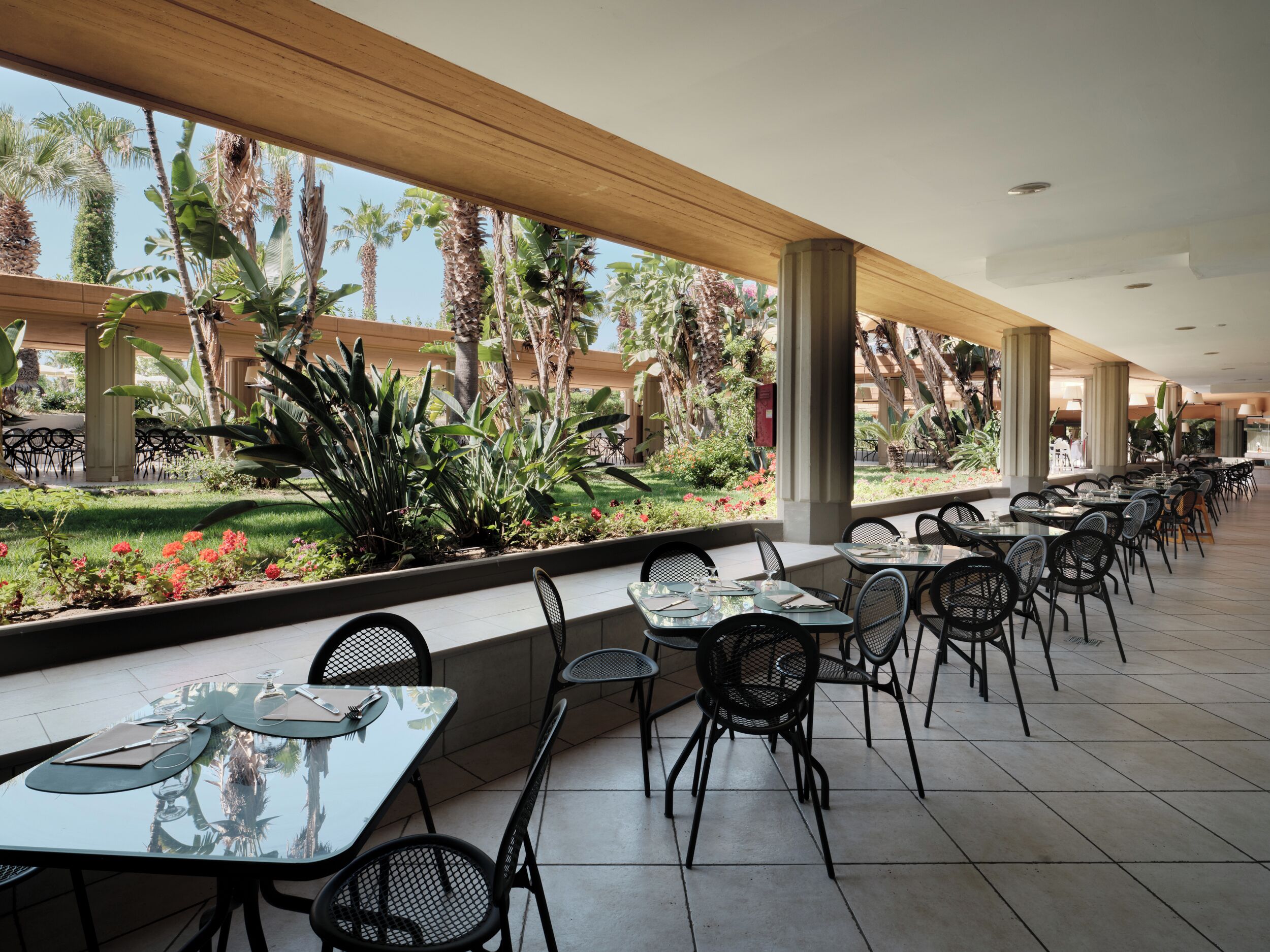 A spacious outdoor dining area with multiple tables and chairs, surrounded by lush greenery and palm trees.