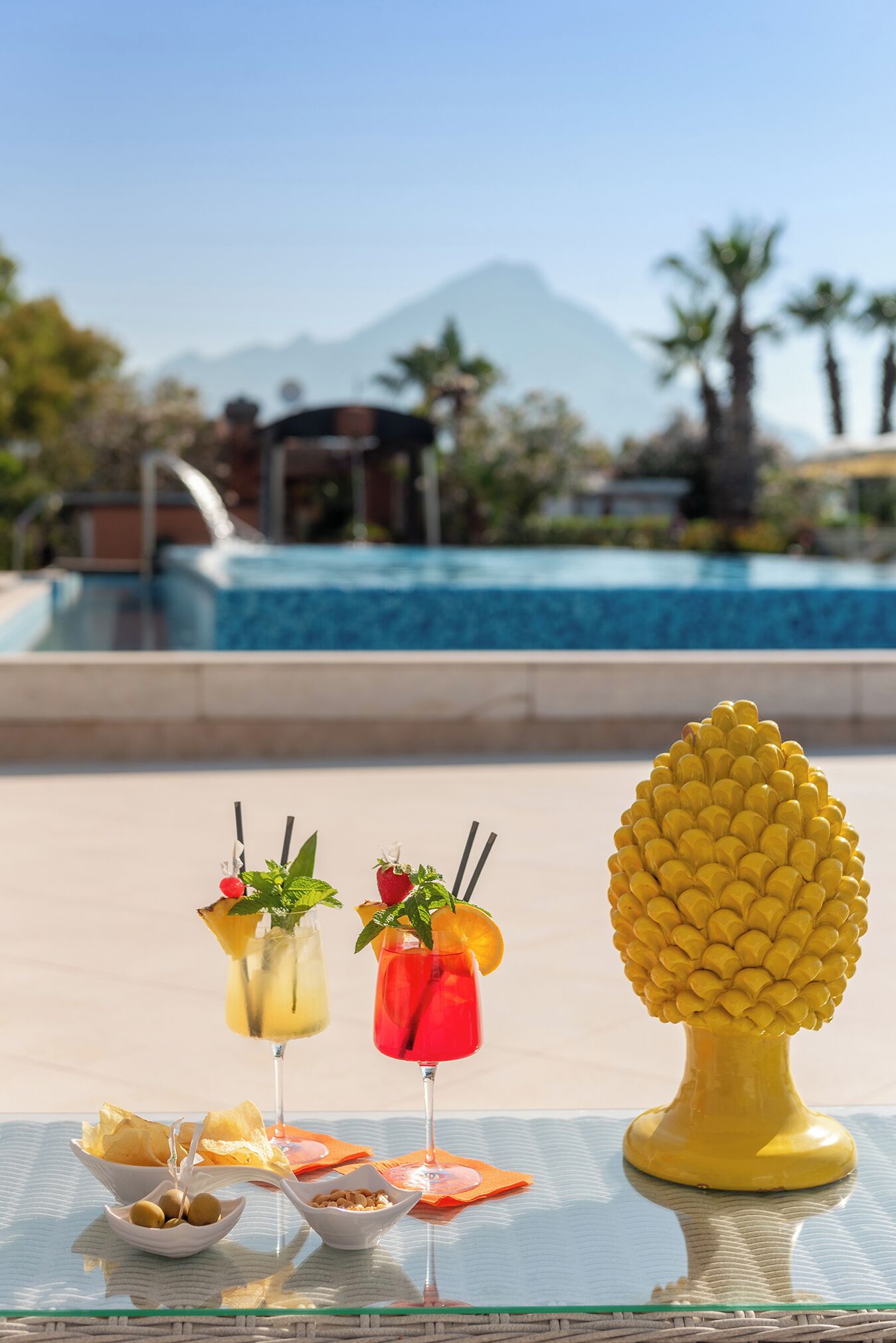 Une table au bord de la piscine, avec des boissons et des en-cas, une sculpture d’ananas décorative et une vue sur les montagnes en arrière-plan.