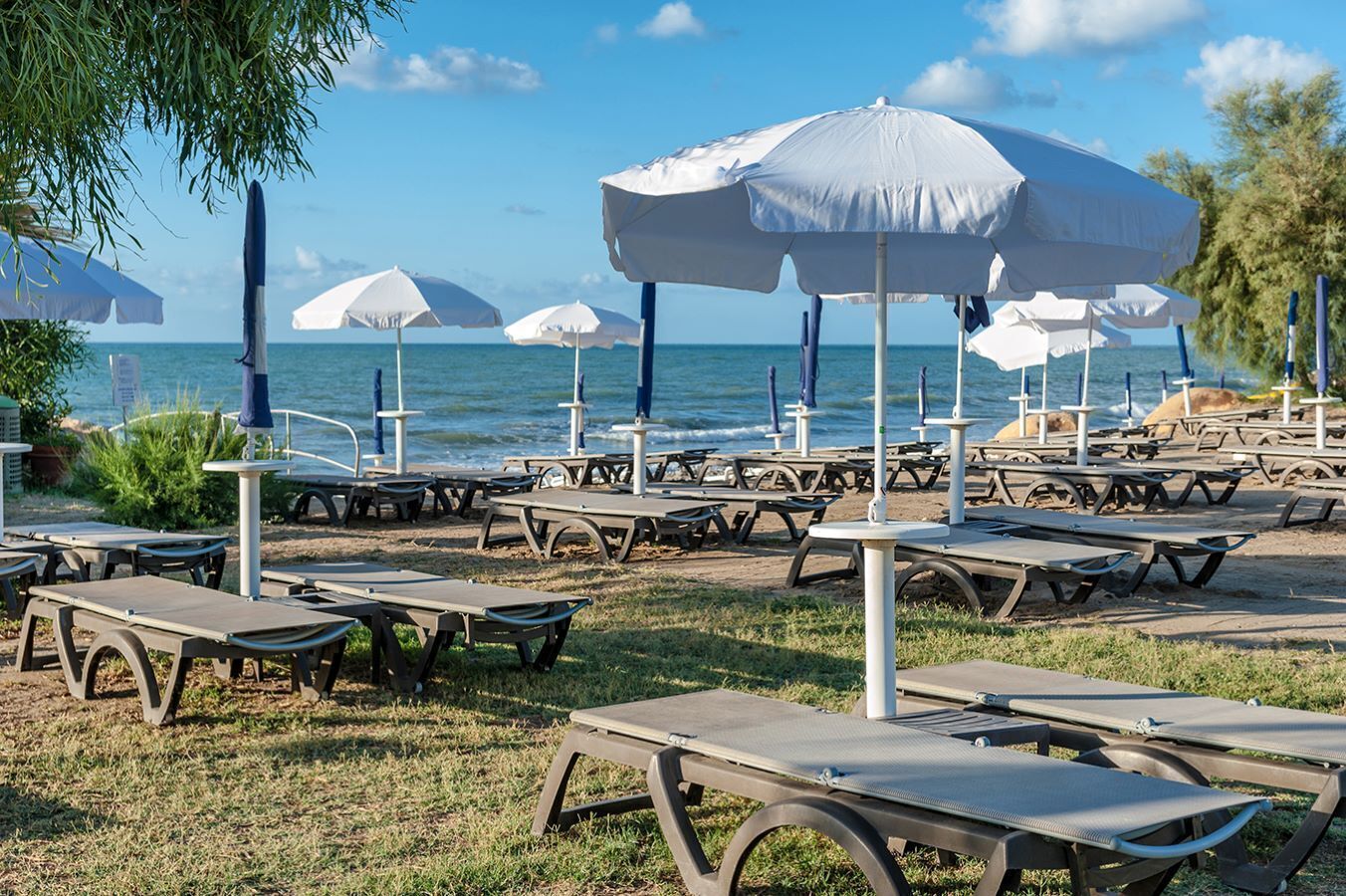 A beachside area with multiple picnic tables and umbrellas, some with blue poles, and a view of the sea in the background.
