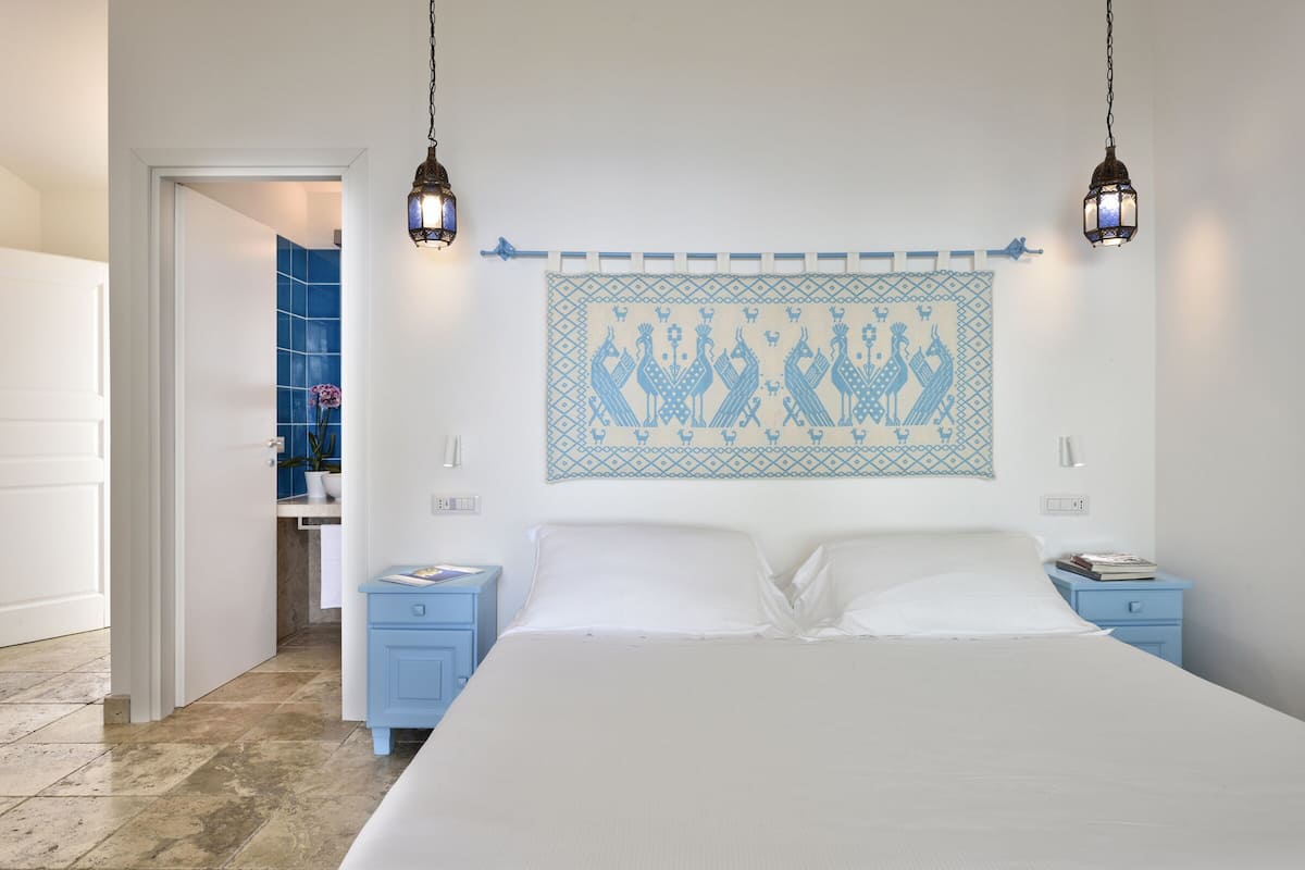 Une chambre à coucher avec un grand lit, deux tables de chevet, une œuvre d’art accrochée au mur et une salle de bain visible à travers une porte ouverte.