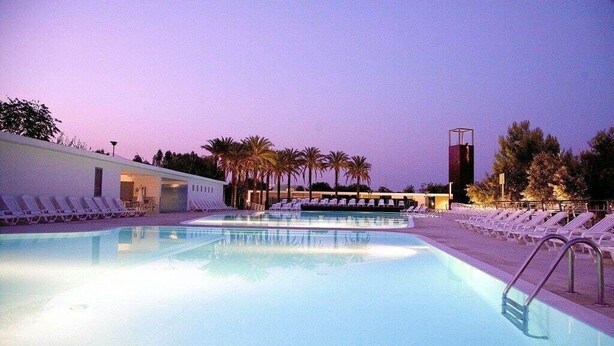 A pool area with lounge chairs and palm trees, surrounded by buildings. - Magna Grecia Village (METAPONTO)