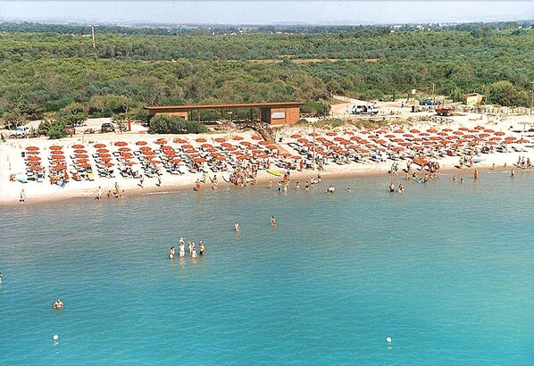 A beach with numerous umbrellas and people swimming. There is a building with a roof and a signboard. - Magna Grecia Village (METAPONTO)