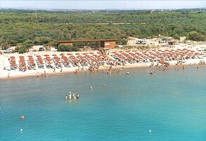 Una playa con numerosos sombrillas y gente nadando. Se observa un edificio con techo y un cartel.