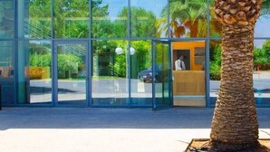 A modern building with large glass windows, a palm tree in front, and a person inside. - Magna Grecia Village (METAPONTO)