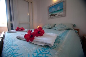 A neatly made bed with a blue and white patterned bedspread, red flowers, and a painting of a beach scene on the wall.