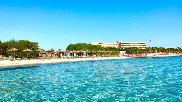 A beach resort with a large building, clear blue water, and umbrellas along the shore. - Club Esse Roccaruja (Stintino)