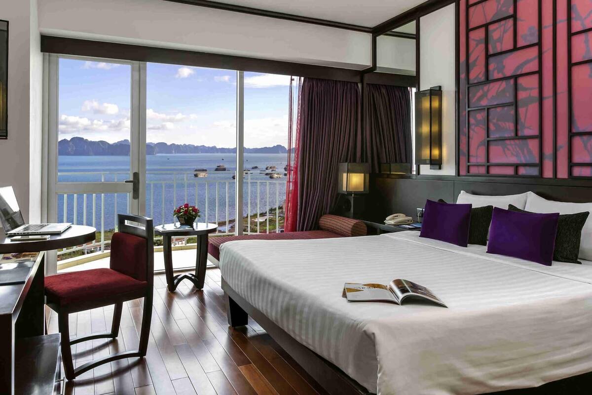 Ein Hotelzimmer mit einem großen Bett, einem Schreibtisch mit einem Laptop, einem Stuhl und Meerblick.