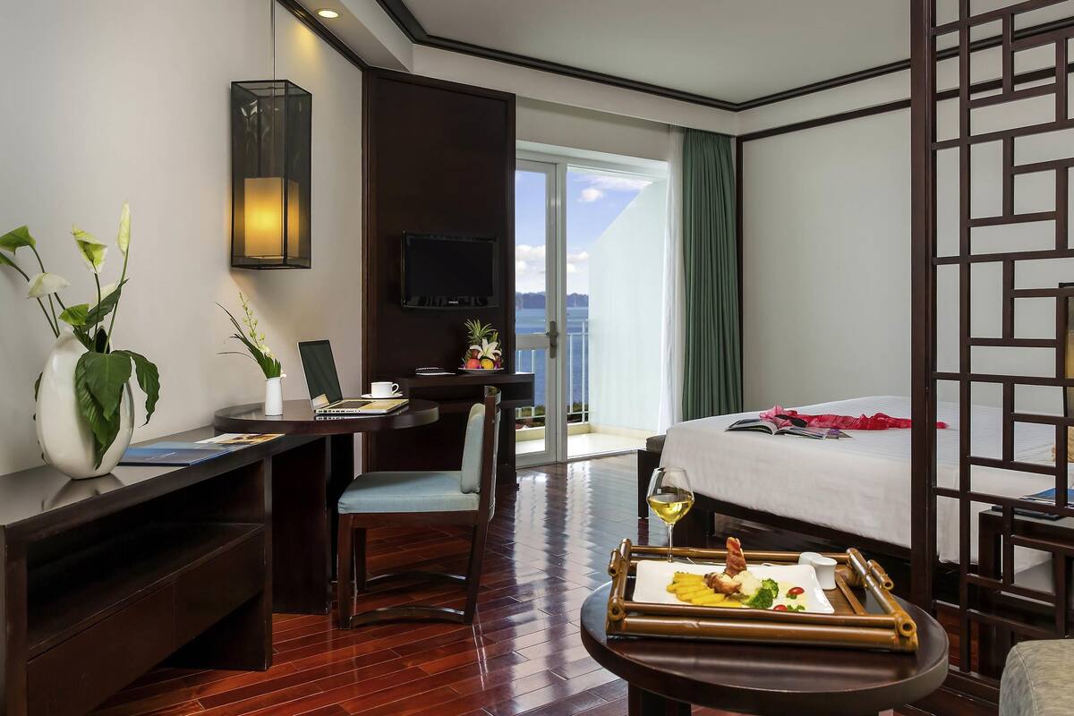 Ein Hotelzimmer mit Bett, Schreibtisch mit Laptop, Esstisch gedeckt mit Speisen, Fernseher an der Wand und Blick aufs Meer.