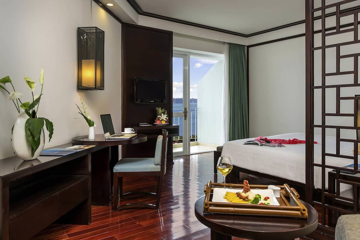 Habitación de hotel con cama, escritorio con computadora portátil, mesa de comedor con comida, televisor en la pared y vista al mar.