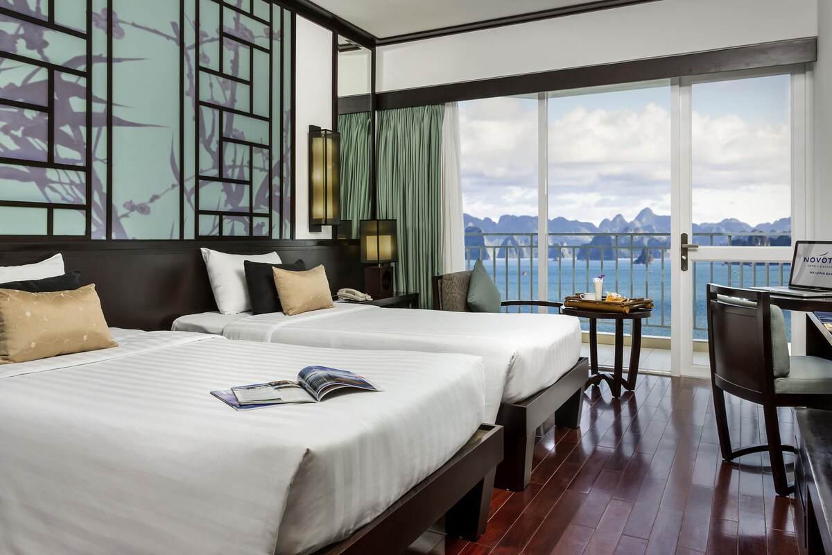 Habitación de hotel con dos camas, un escritorio con una computadora portátil, vista al mar y un balcón con barandilla.