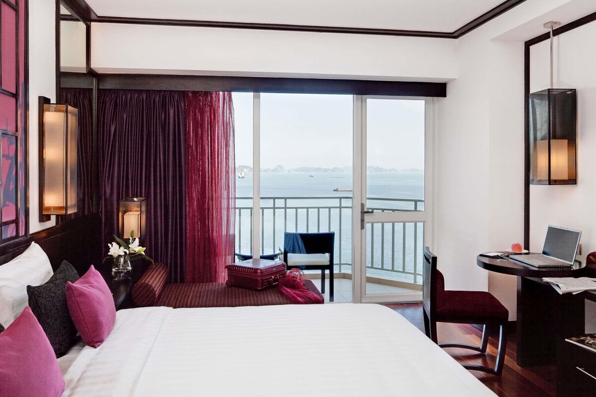 Habitación de hotel con una cama grande, un escritorio con una computadora portátil, una silla, una mesita con un jarrón de flores y vista al mar a través de un ventanal amplio.