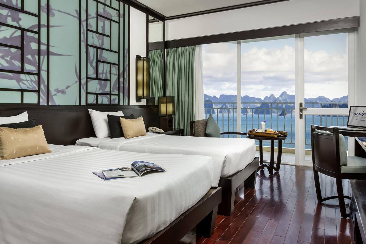 Ein Hotelzimmer mit zwei Betten, einem Schreibtisch mit einem Laptop, Meerblick und einem Balkon mit Geländer.