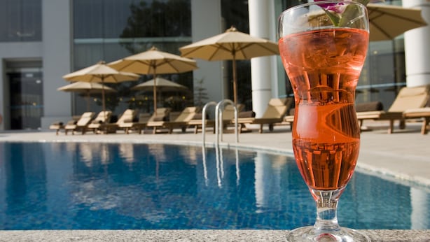 A clear glass of a pink drink with a flower garnish, placed on a poolside with lounge chairs and umbrellas in the background.