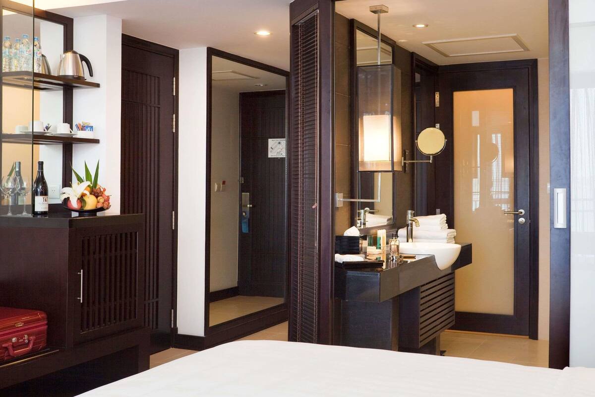 A hotel room with a bed, a bathroom with a sink and mirror, a minibar, and a red suitcase.