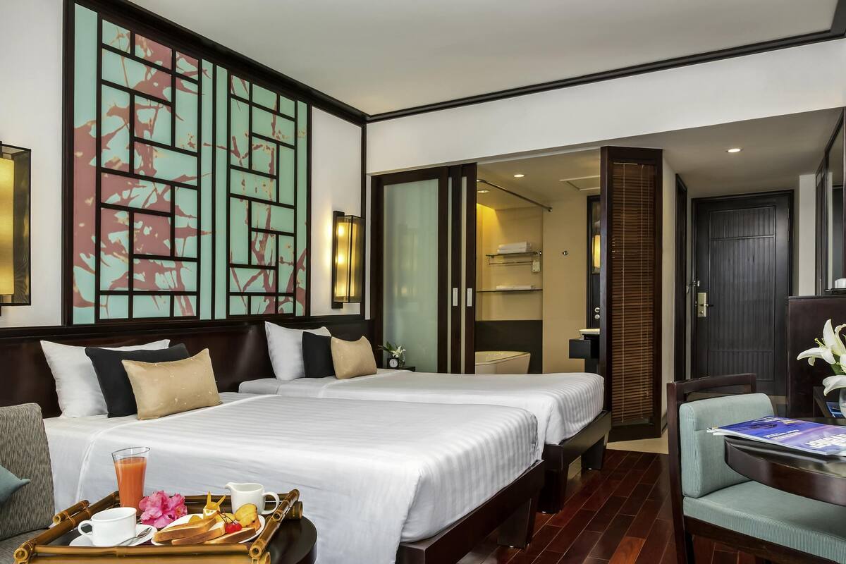 A hotel room with two beds, a dining area, a bathroom, and a view of the city.
