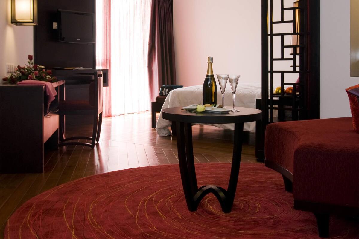A hotel room with a bed, a round table with a bottle and glasses, a red rug, and a TV on the wall.