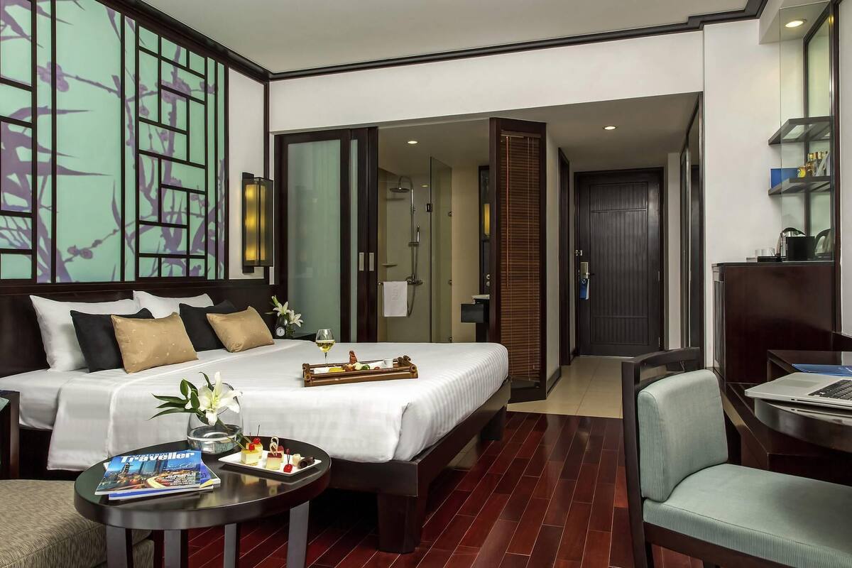 A hotel room with a large bed, a desk, a chair, a small table with a magazine, and a bathroom visible through an open door.