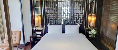 A hotel room with a large bed, a chair, a nightstand with a lamp, and a decorative wall panel.