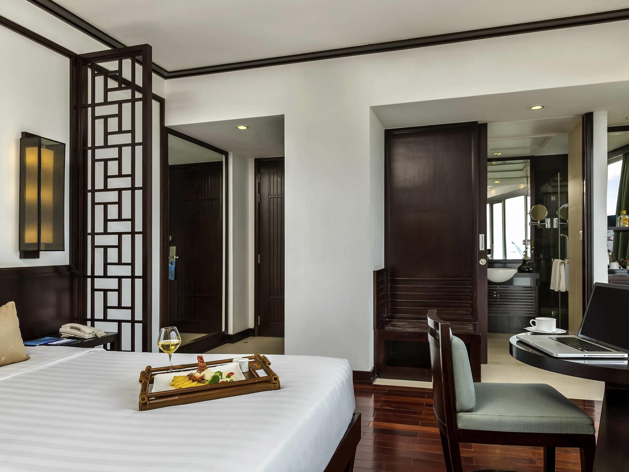 Habitación de hotel con una cama, un servicio de comidas, una silla, un escritorio con una computadora portátil y un baño visible a través de una puerta abierta.