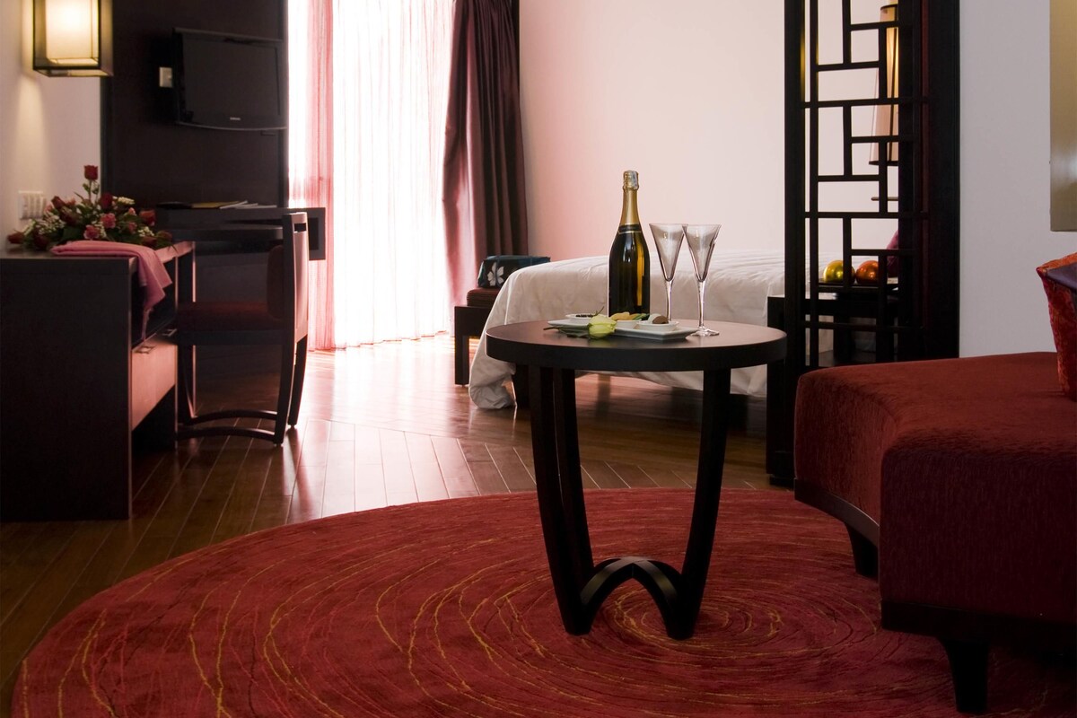 Habitación de hotel con una cama, una mesa redonda con una botella y vasos, una alfombra roja y un televisor en la pared.