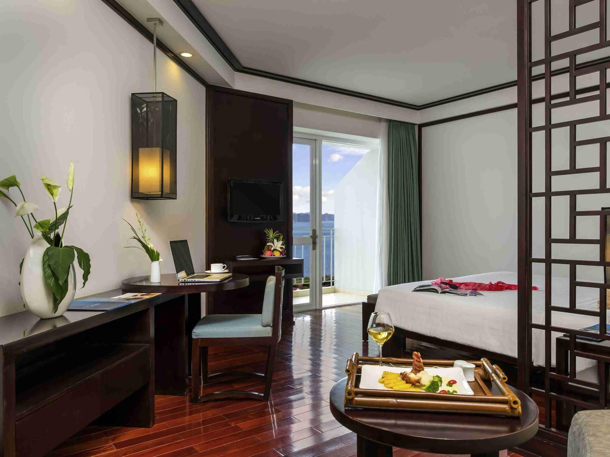 Habitación de hotel con cama, escritorio con una computadora portátil, mesa de comedor con comida y televisor en la pared.