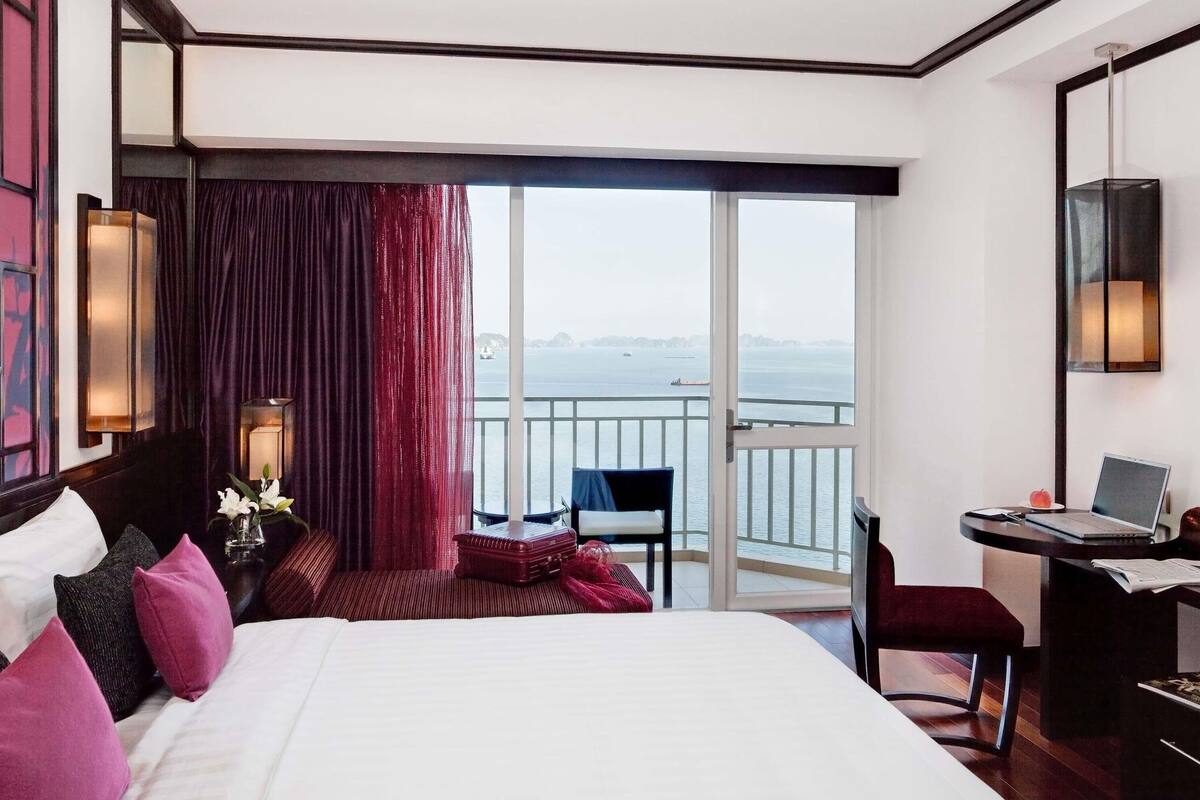 A hotel room with a large bed, a desk with a laptop, a chair, a small table with a vase of flowers, and a view of the sea through a large window.