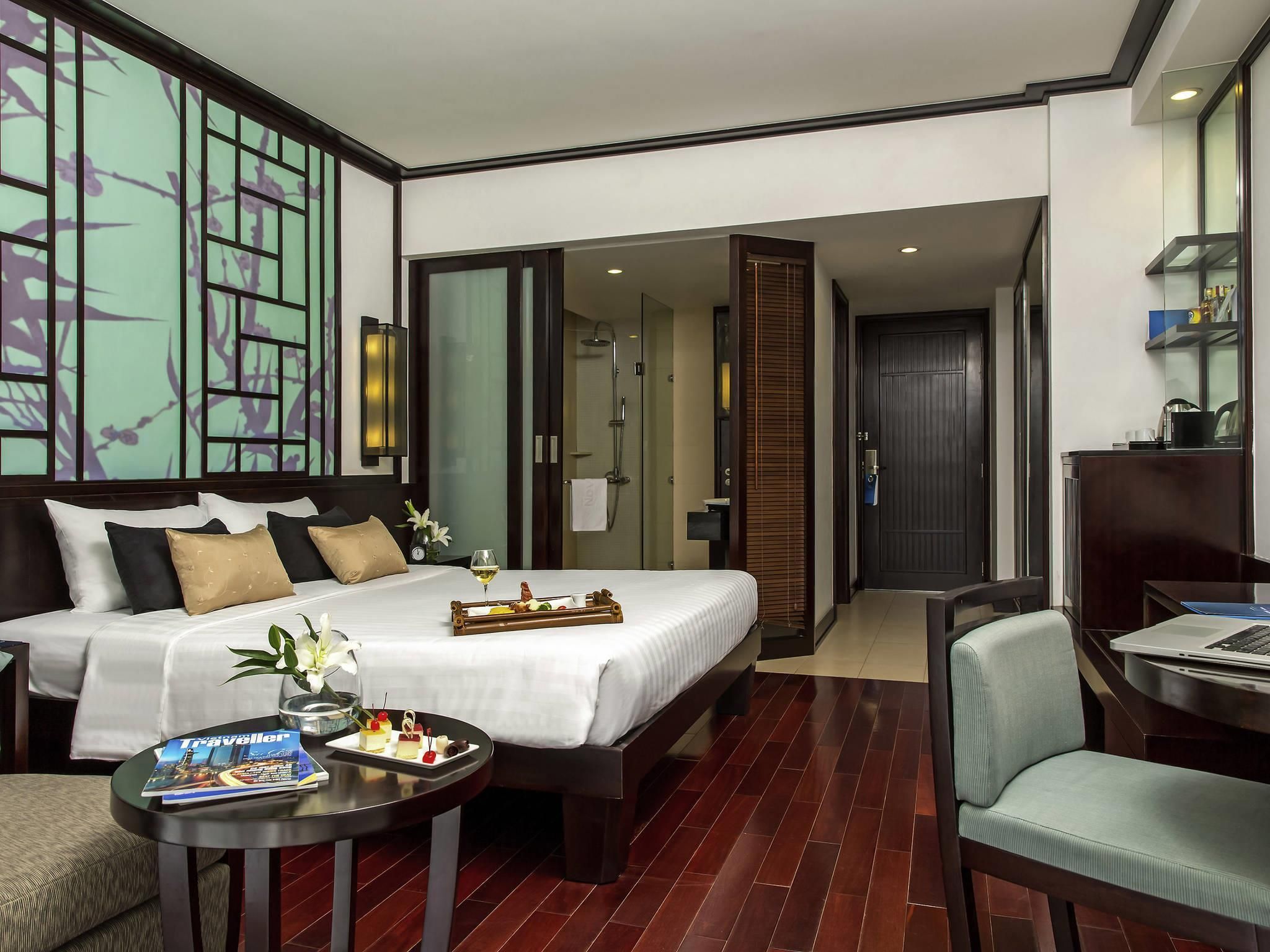 Habitación de hotel con una cama grande, un escritorio, una silla, una mesita con una revista y un baño visible a través de una puerta abierta.