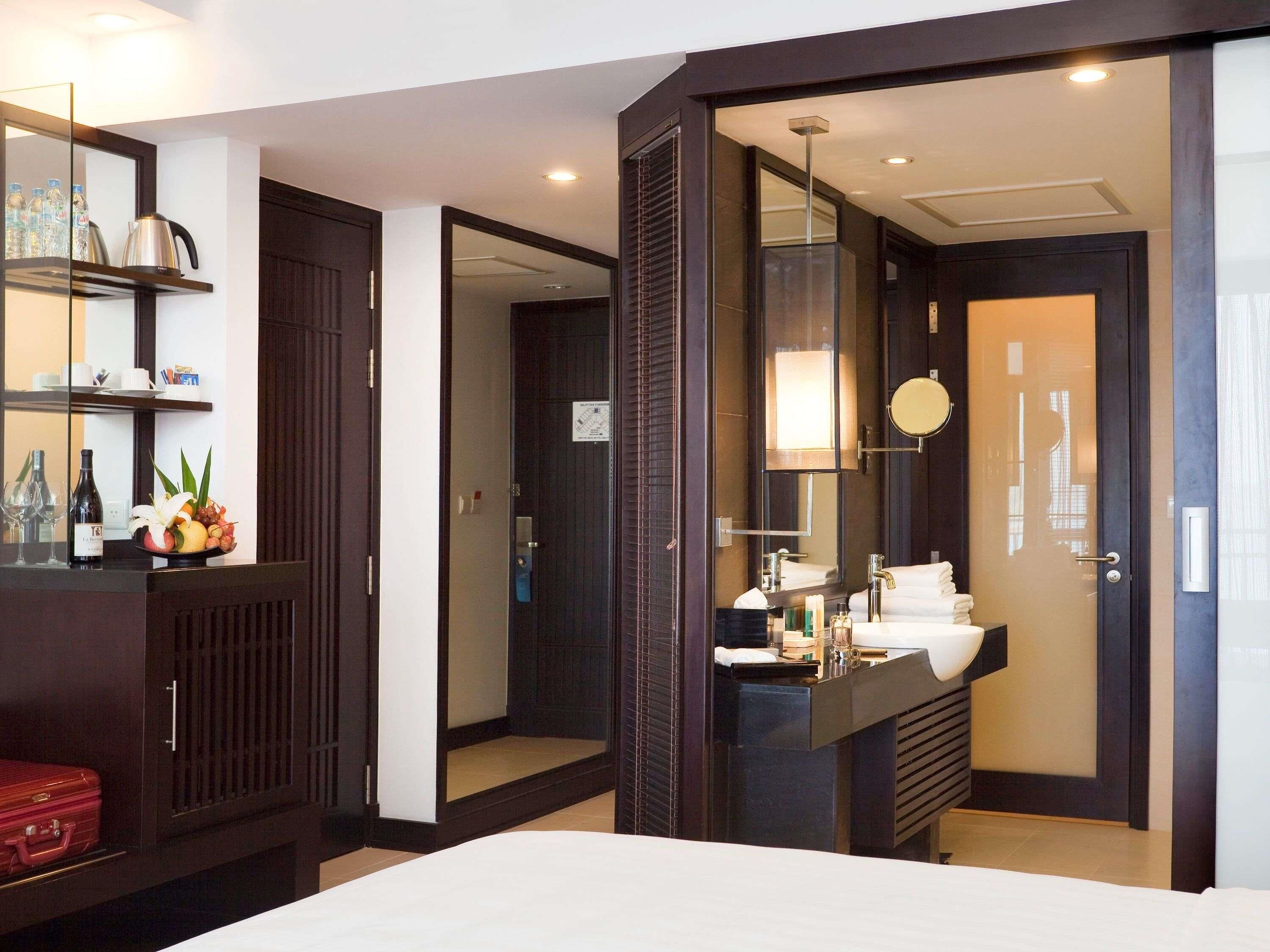 Une chambre d’hôtel avec un lit, une salle de bain comprenant un lavabo et un miroir, un mini-bar et une valise rouge.