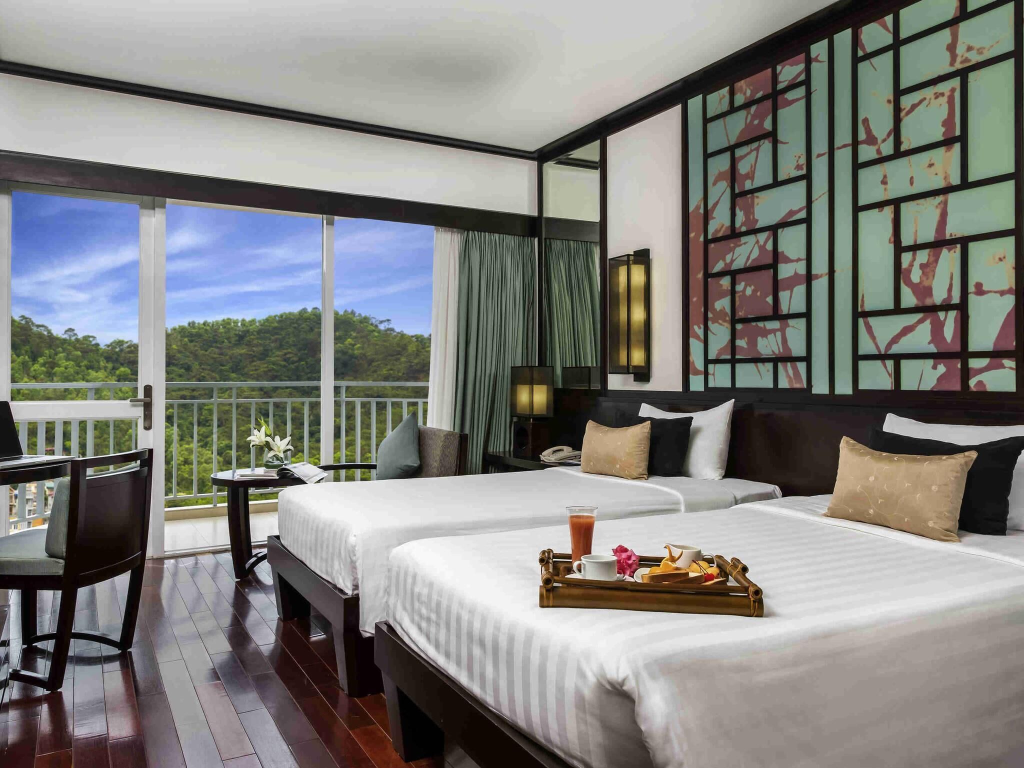 Habitación de hotel con dos camas, un escritorio, un balcón con vistas al bosque y un adorno en la pared.