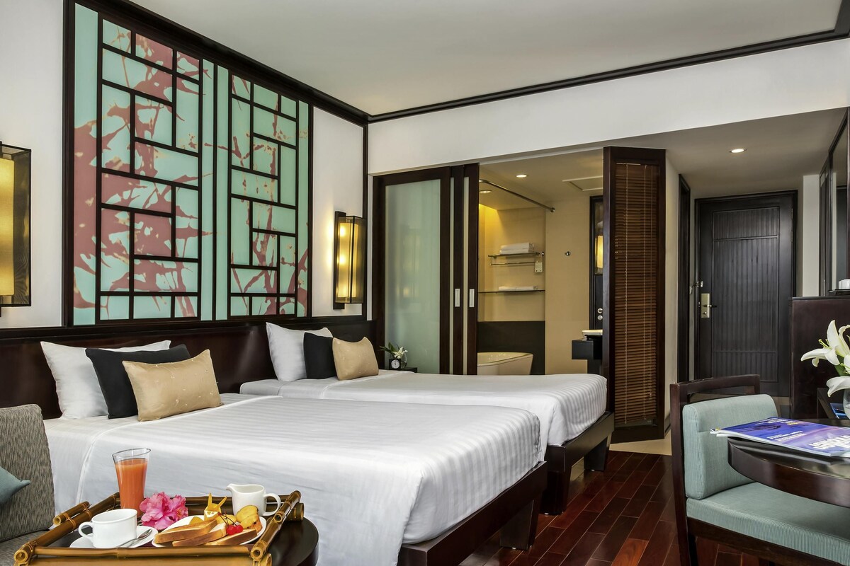 Habitación de hotel con dos camas, zona de comedor, baño y vistas a la ciudad.