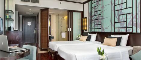 A hotel room with a large bed, a desk with a laptop, a chair, and a small table with a book and a vase of flowers.