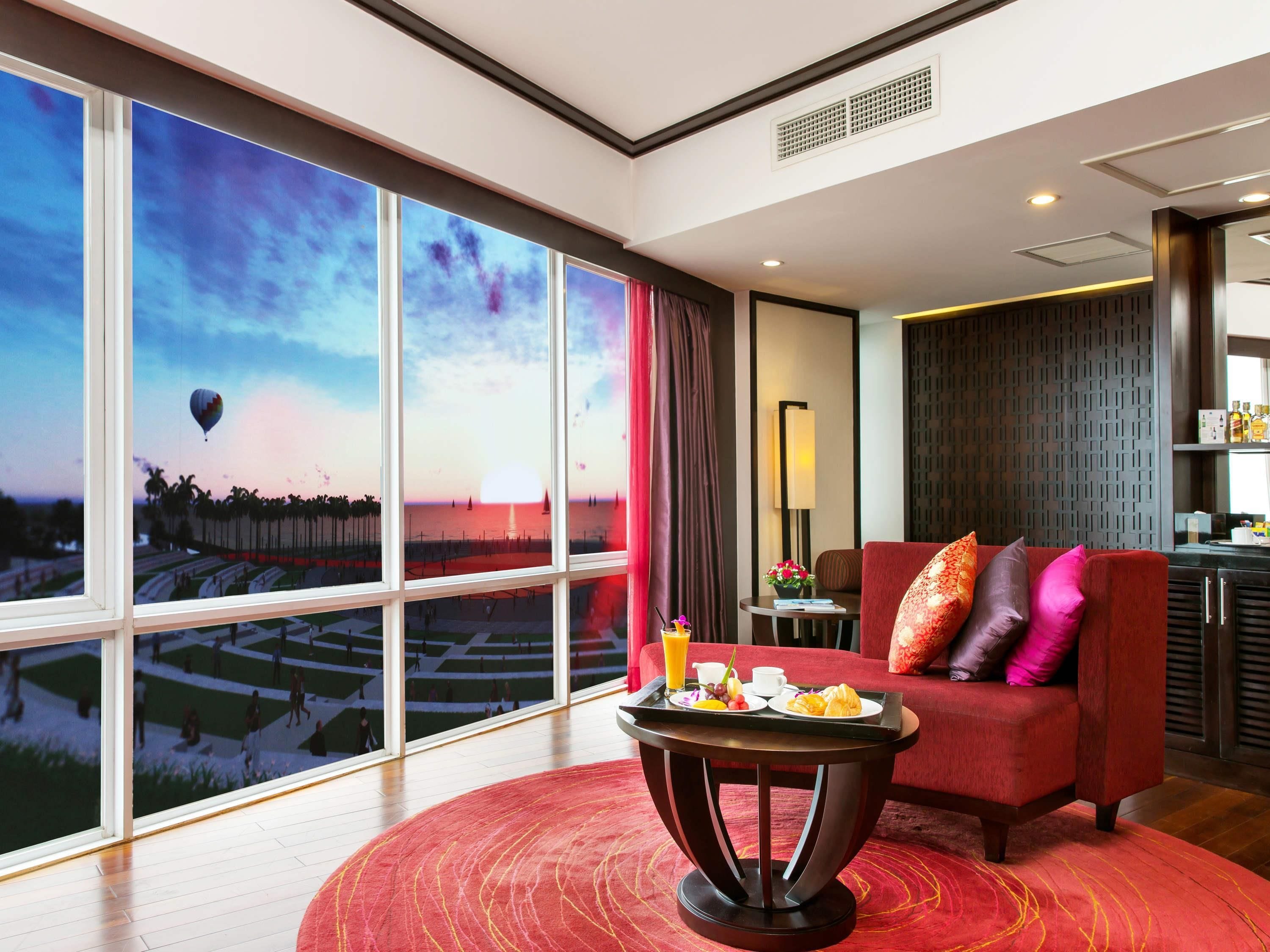 Ein Hotelzimmer mit großem Fenster und Blick auf einen Sonnenuntergang über dem Strand, einem roten Sofa, einem kleinen Tisch mit Frühstücksangeboten und einem modernen Interieur.