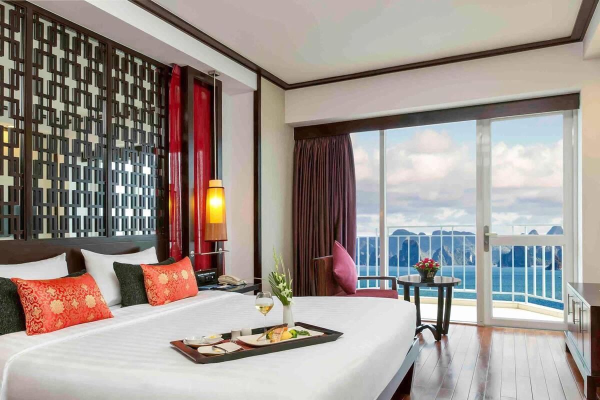 Ein Hotelzimmer mit einem großen Bett, einem Esstisch mit einer Servitorenplatte voller Speisen, einer Lampe, einem Stuhl und Blick auf das Meer.