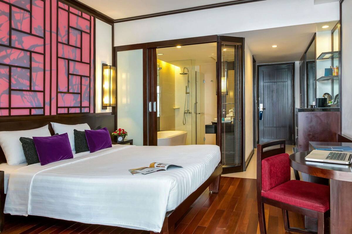 Ein Hotelzimmer mit einem Bett, einem Schreibtisch mit einem Laptop, einer Duschkabine und einer Glastür zum Badezimmer.