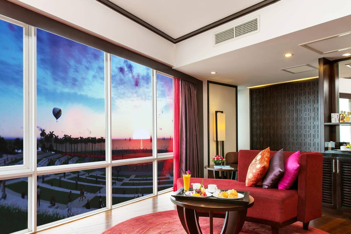 A hotel room with a large window offering a view of a sunset over a beach, a red sofa, a small table with breakfast items, and a modern interior design.
