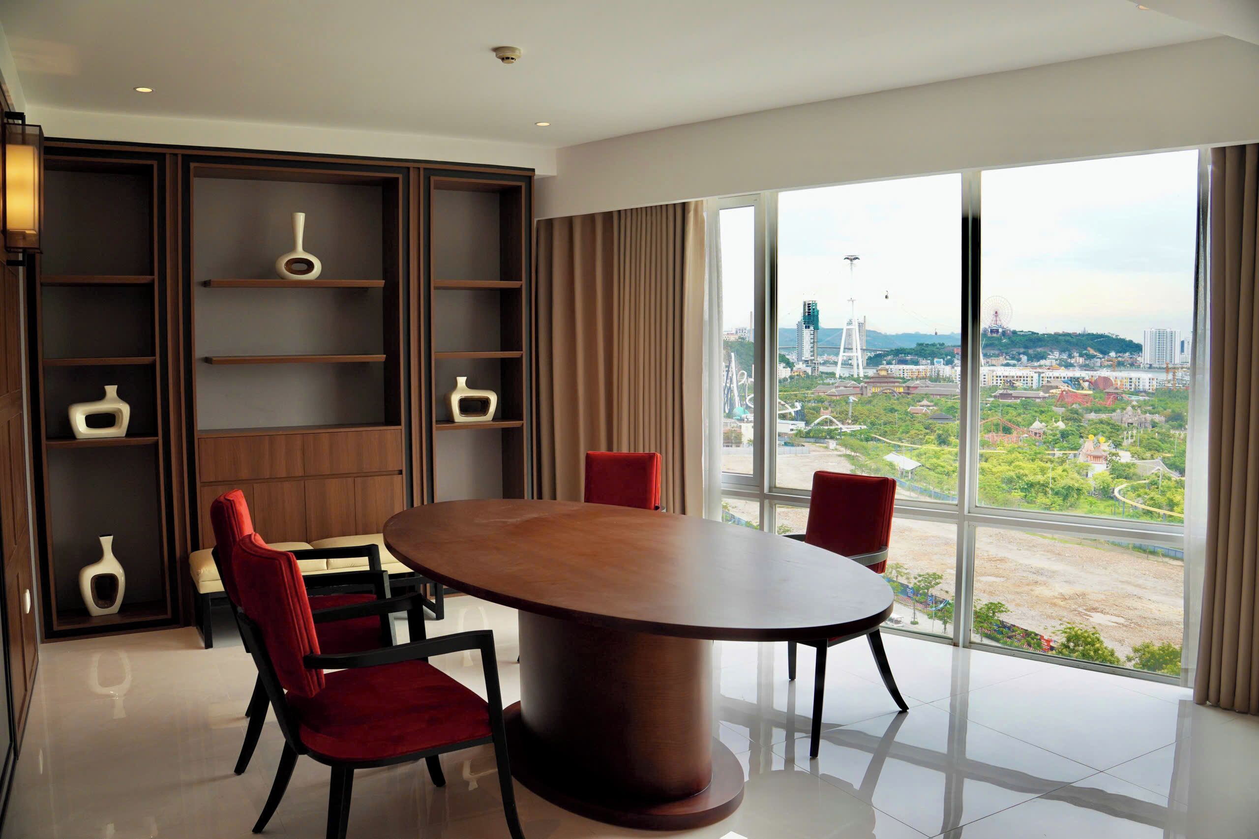 A modern hotel room with a large round table, red chairs, a built-in bookshelf, and a large window offering a city view.
